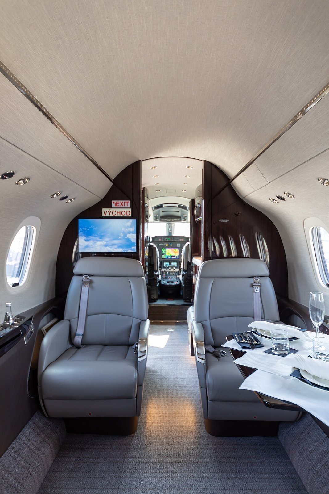 Forward cabin of Citation Longitude OK-JRE at EBACE 2023, showing grey seats, wood trim and a set dining table.